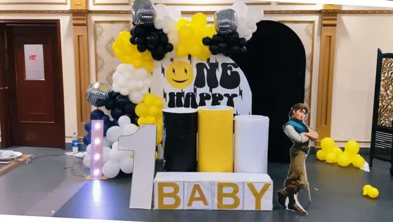 King Flowers A decorated party setup with a ONE HAPPY sign, clusters of yellow, white, and black balloons, disco balls, a cutout of a cartoon character, and big 1 and BABY blocks in front, for a first birthday celebration.
